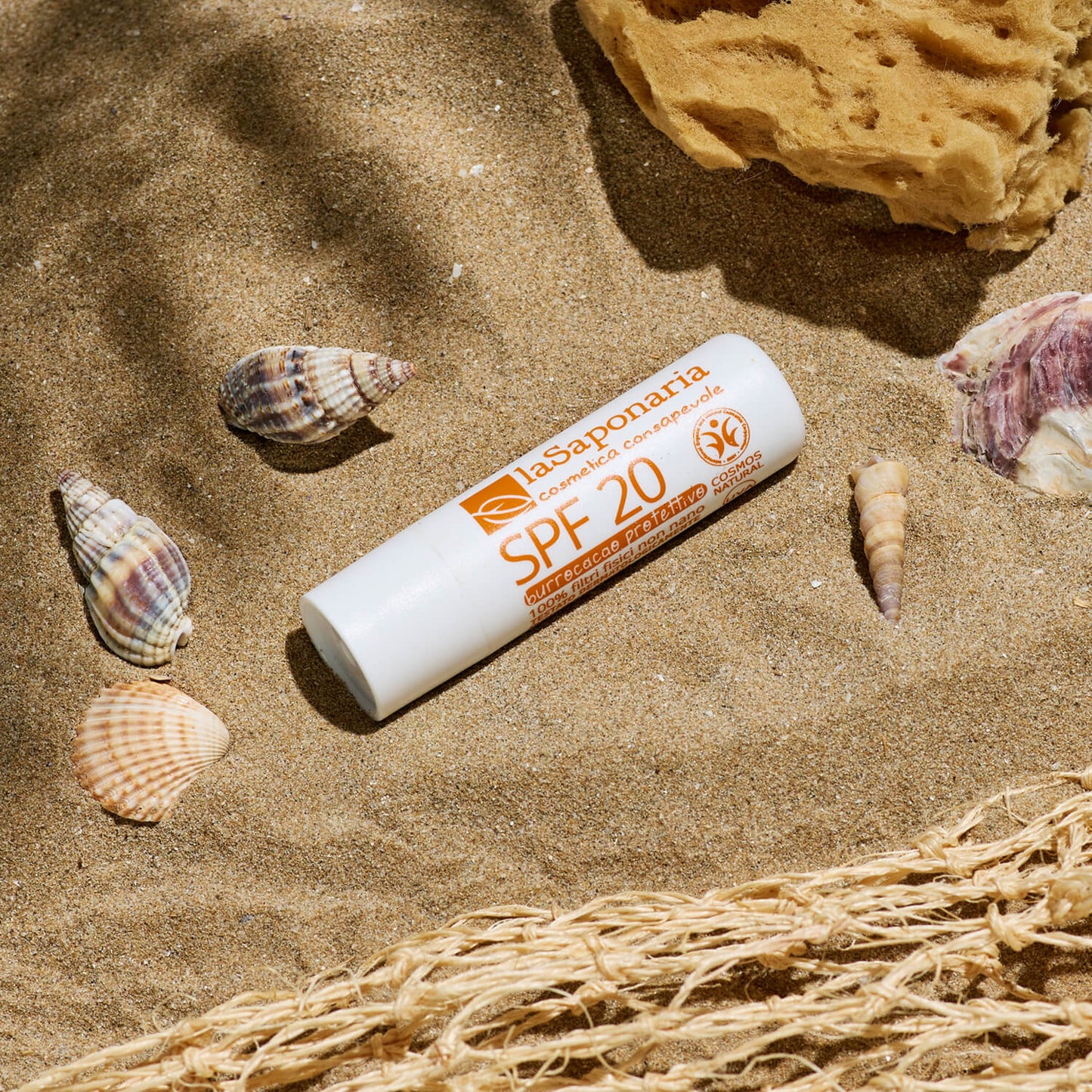 Bálsamo Labial Protetor SPF 20 - Biocao da La Saponaria na areia de uma praia com conchas